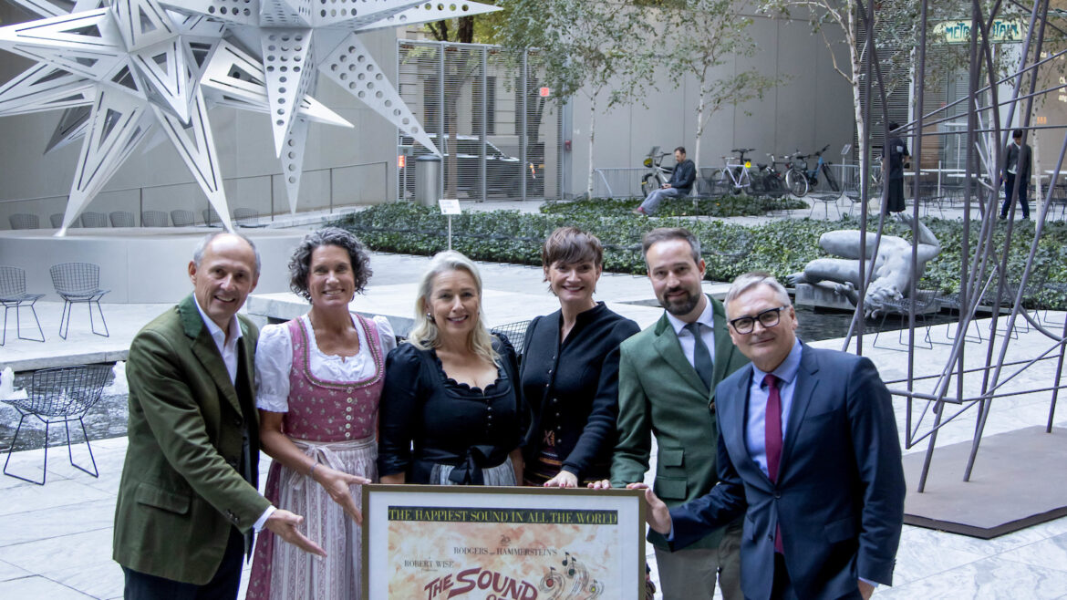 Sechs Personen posieren im Freien mit einem "The Sound of Music"-Plakat; große Sternenskulpturen im Hintergrund.