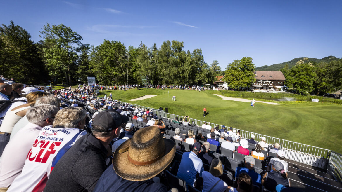 Die Zuschauer beobachten die Golfer auf einem grünen Golfplatz unter blauem Himmel, umgeben von Bäumen und Gebäuden.