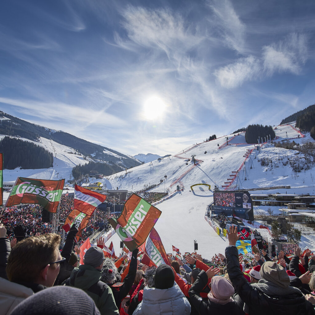 Fahnenschwenkende Menschenmenge bei einer verschneiten Skiveranstaltung mit Bergen und einer strahlenden Sonne im Hintergrund.