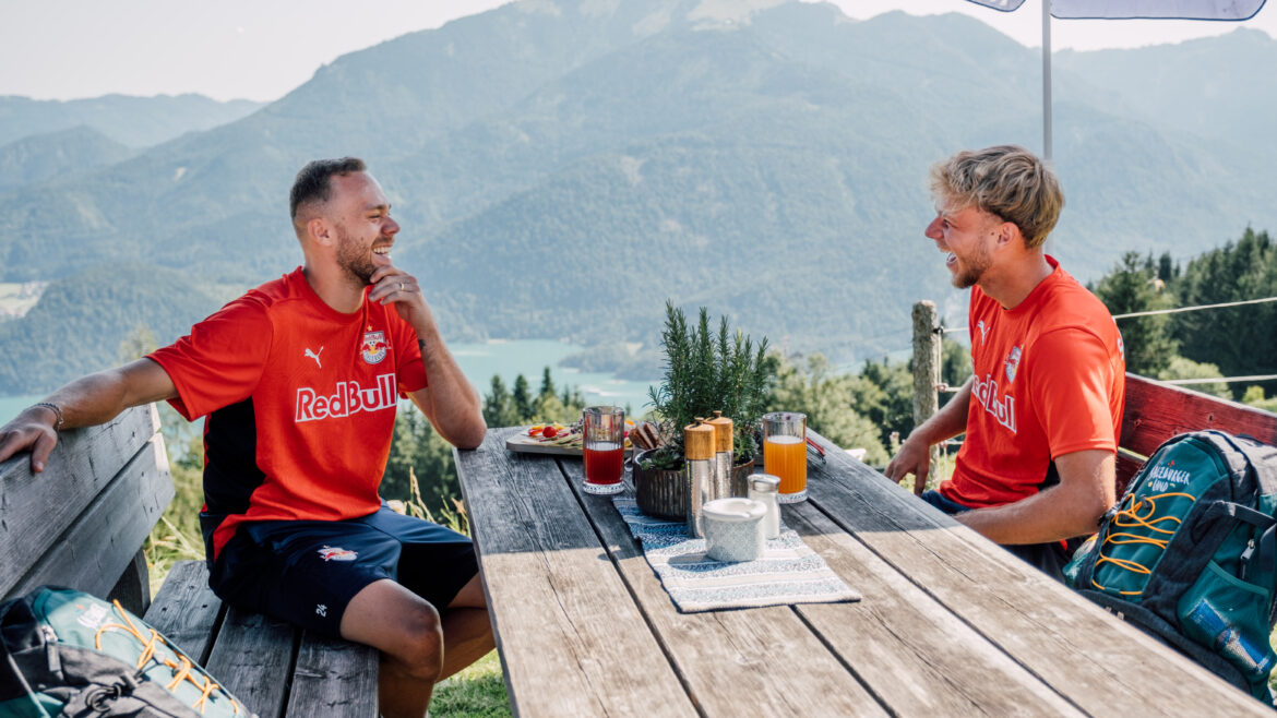 Zwei Männer in Red Bull-Hemden sitzen an einem Picknicktisch im Freien und lachen, während im Hintergrund die Berge zu sehen sind.