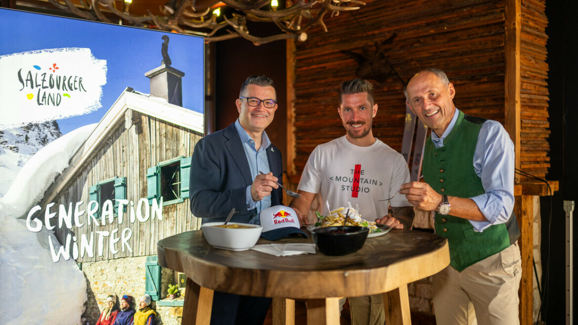 Drei Männer lächeln und genießen das Essen an einem rustikalen Tisch, mit einem Wintertourismusplakat im Hintergrund.