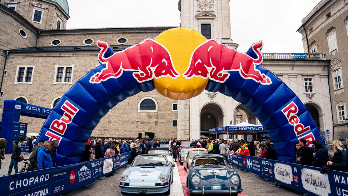Zwei Oldtimer fahren bei einem Auto-Event unter einem Red-Bull-Bogen hindurch, mit Menschenmengen und historischen Gebäuden im Hintergrund.