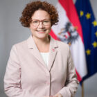 Eine Frau in einem hellrosa Blazer steht lächelnd vor einer österreichischen und einer EU-Flagge.