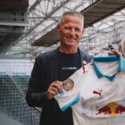 Ein lächelnder Mann in einem Blazer hält ein von Puma Red Bull gesponsertes Fußballtrikot in einem Stadion hoch.