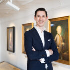 Ein lächelnder Mann in einem marineblauen Anzug steht mit verschränkten Armen in einer Kunstgalerie mit gerahmten Porträts an der Wand.