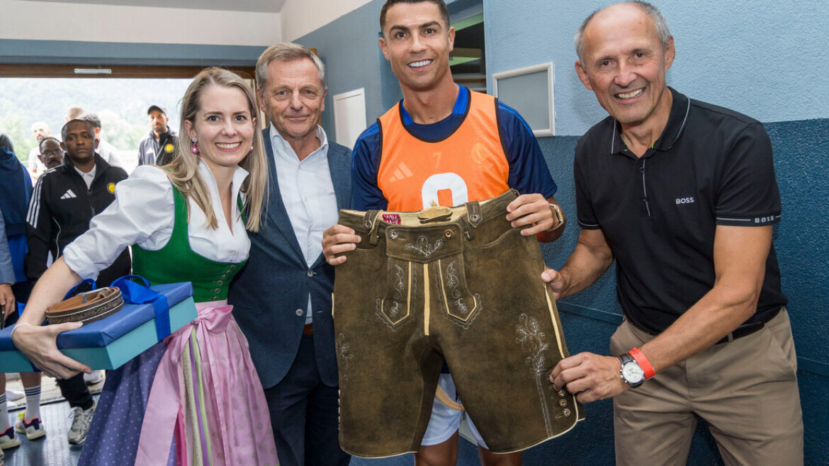 Vier Personen lächeln; eine hält eine traditionelle österreichische Lederhose und eine andere hält eingepackte Geschenke in einem Flur.