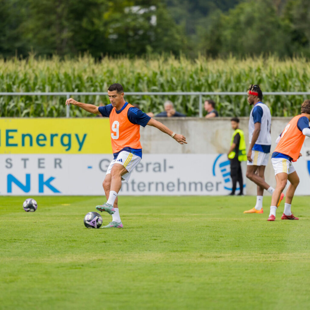 Fußballspieler in orangefarbenen Westen üben auf einer Wiese in der Nähe eines Maisfeldes und von Werbetafeln.