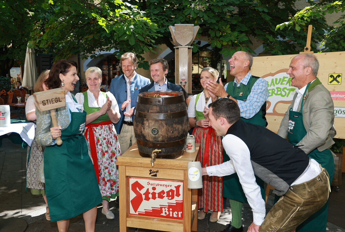Eine Gruppe in traditioneller Kleidung versammelt sich bei einem Fest im Freien um ein Bierfass, lächelt und lacht.
