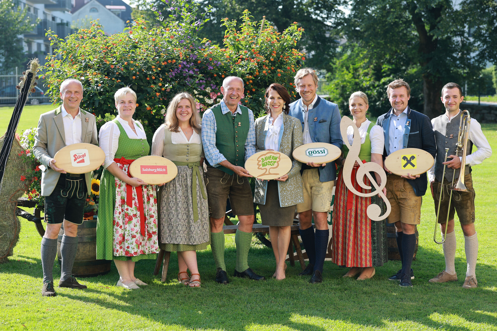 Acht Personen in traditioneller Kleidung stehen im Freien, lächeln und halten Schilder mit Logos und Musiksymbolen.