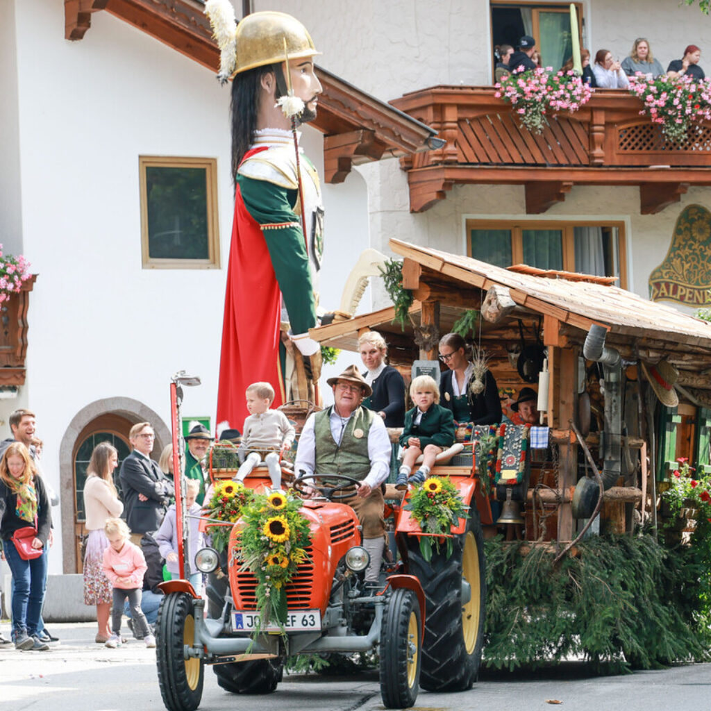 Ein Umzugswagen mit einer riesigen Figur und Menschen in österreichischer Tracht, die auf einem mit Sonnenblumen geschmückten Traktor fahren.