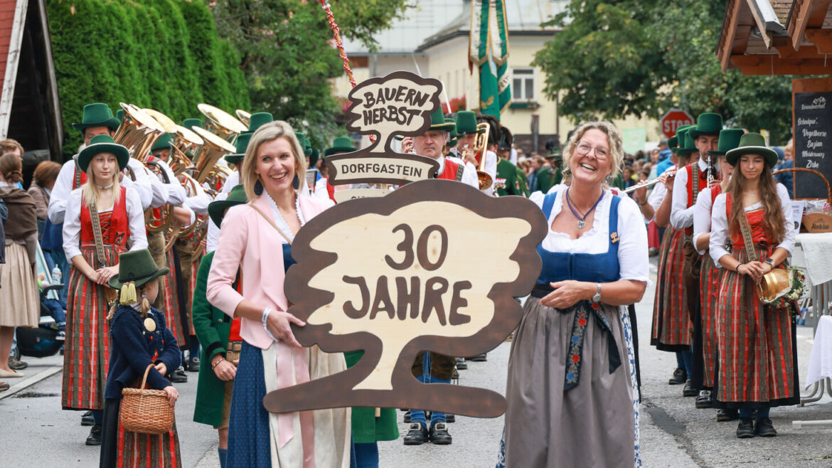 Zwei Frauen in traditioneller Kleidung halten ein "30 Jahre"-Schild bei einer festlichen Parade mit einer Musikkapelle und einer Kirche im Hintergrund.