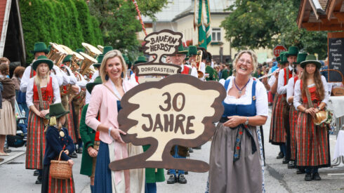 Zwei Frauen in traditioneller Kleidung halten ein "30 Jahre"-Schild bei einer festlichen Parade mit einer Musikkapelle und einer Kirche im Hintergrund.