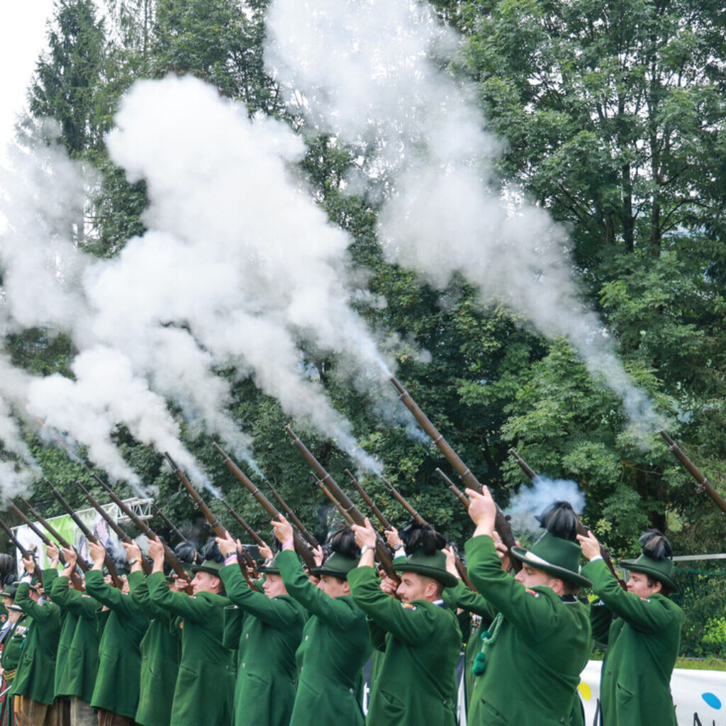Eine Gruppe von Personen in grünen Uniformen schießt in einer baumbestandenen Umgebung Gewehre in die Luft und erzeugt Rauch.