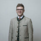 Lächelnder Mann mit Brille und traditioneller beigefarbener geknöpfter Jacke vor einem schlichten hellen Hintergrund.