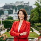 Frau in rotem Blazer, die in einem Garten mit einem historischen Schloss und einer Stadtlandschaft im Hintergrund steht.