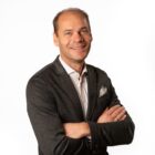 Ein lächelnder Mann in einem dunklen Blazer steht mit verschränkten Armen vor einem schlichten weißen Hintergrund.