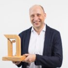 Ein lächelnder Mann in einem Blazer hält eine moderne Holzskulptur vor einem schlichten weißen Hintergrund.