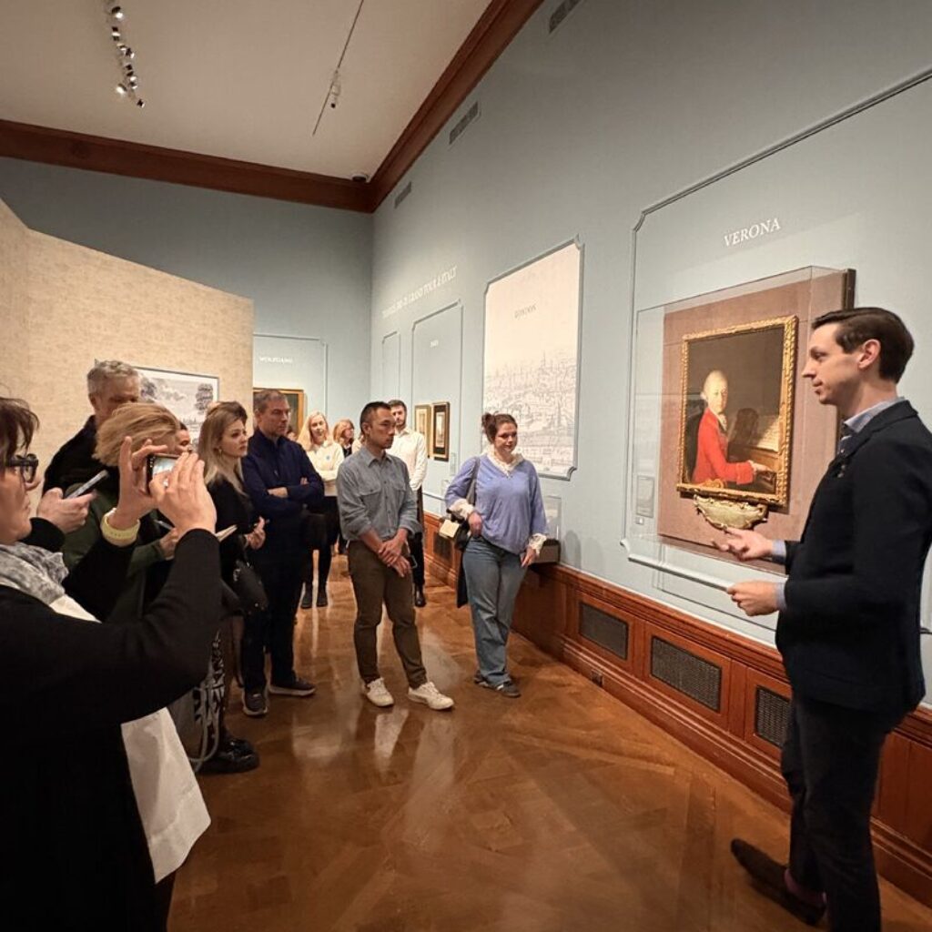 Eine Gruppe von Menschen hört einem Sprecher zu, der neben einem gerahmten Mozart Gemälde an einer Galeriewand spricht.