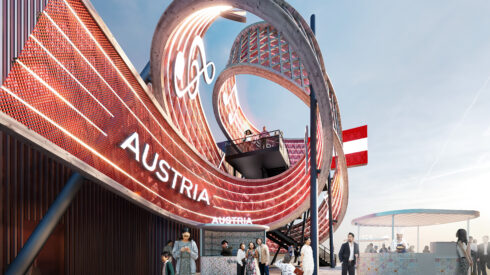 Ein geschwungener, moderner Pavillon mit der Aufschrift „AUSTRIA“, rot leuchtenden Lichtern und Menschen, die in der Nähe vorbeigehen; die österreichische Flagge ist zu sehen.