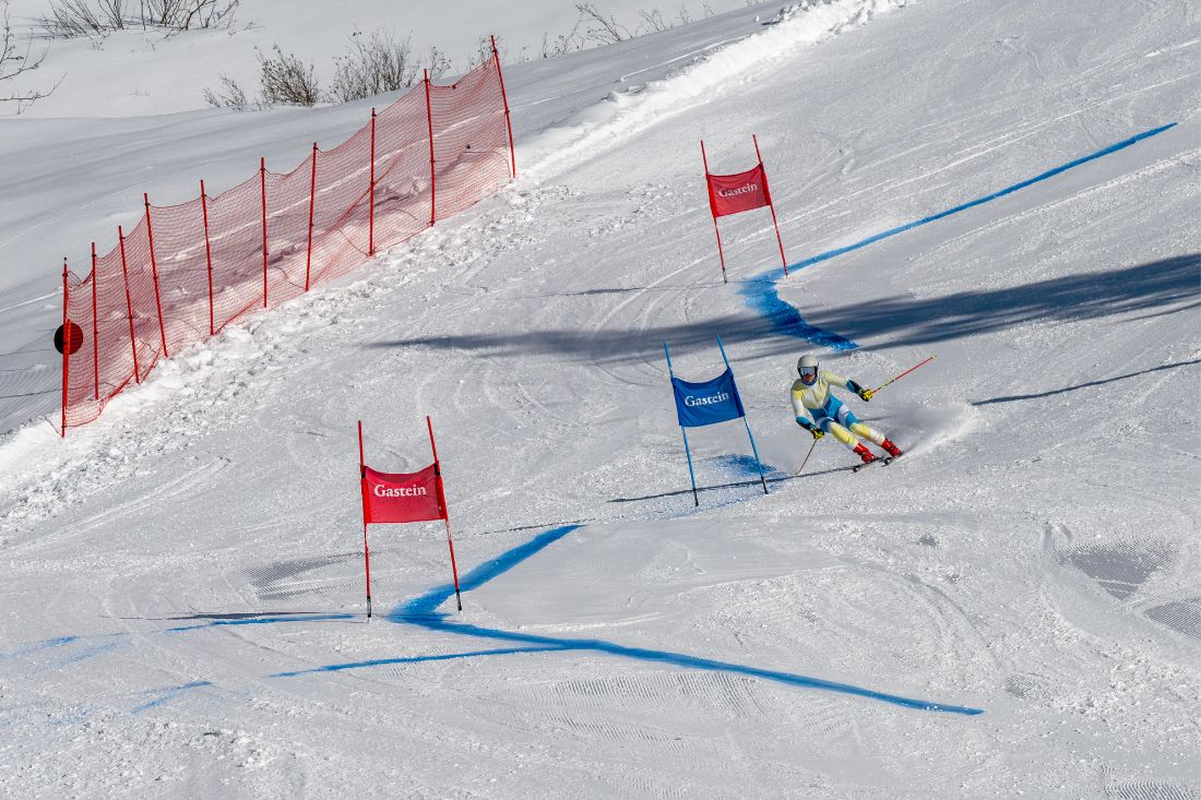 Ein Skifahrer rast einen verschneiten Hang hinunter und passiert dabei rote und blaue Slalomtore neben einem Sicherheitszaun.