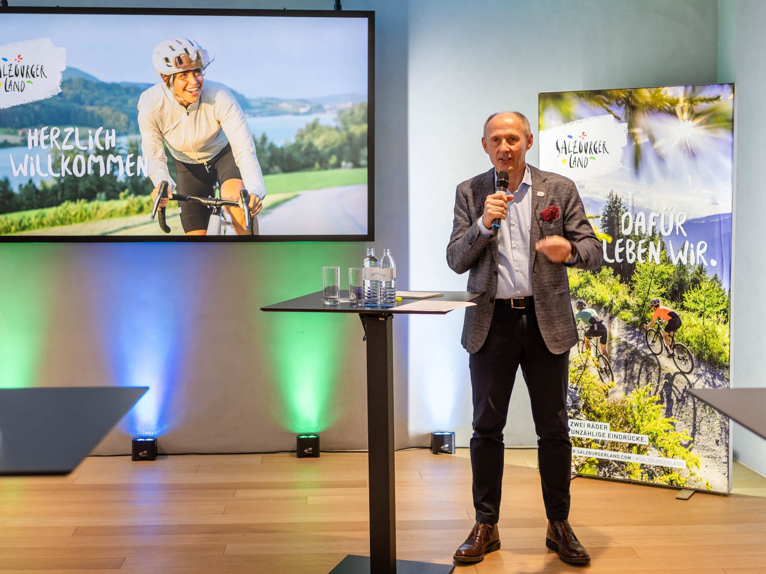 Ein Mann hält eine Rede am Rednerpult; hinter ihm ist auf einer Leinwand ein Radfahrer und der Text „Herzlich Willkommen“ zu sehen.