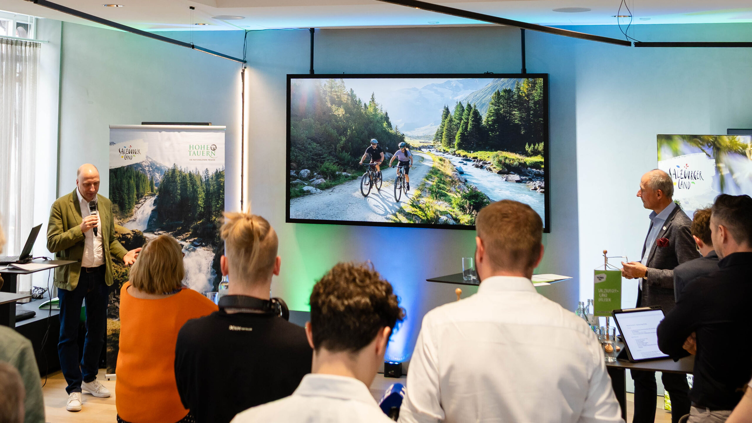 Ein Redner hält vor einem Publikum einen Vortrag, während auf einer großen Leinwand zwei Radfahrer in einer malerischen Berglandschaft zu sehen sind.