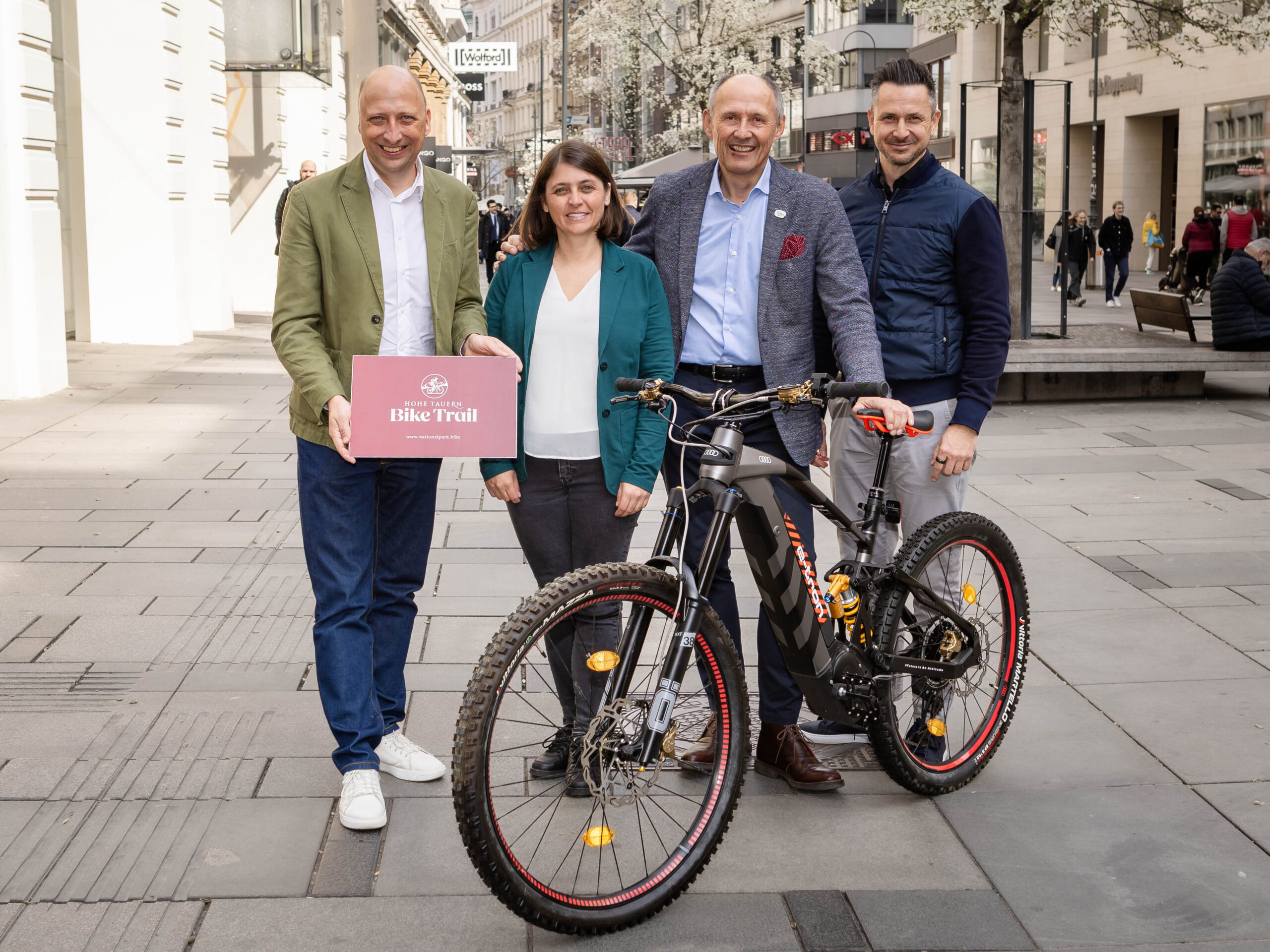 Vier Personen stehen mit einem Mountainbike auf einer Straße in der Stadt; eine von ihnen hält ein Schild mit der Aufschrift „Bike Trail“ in der Hand.