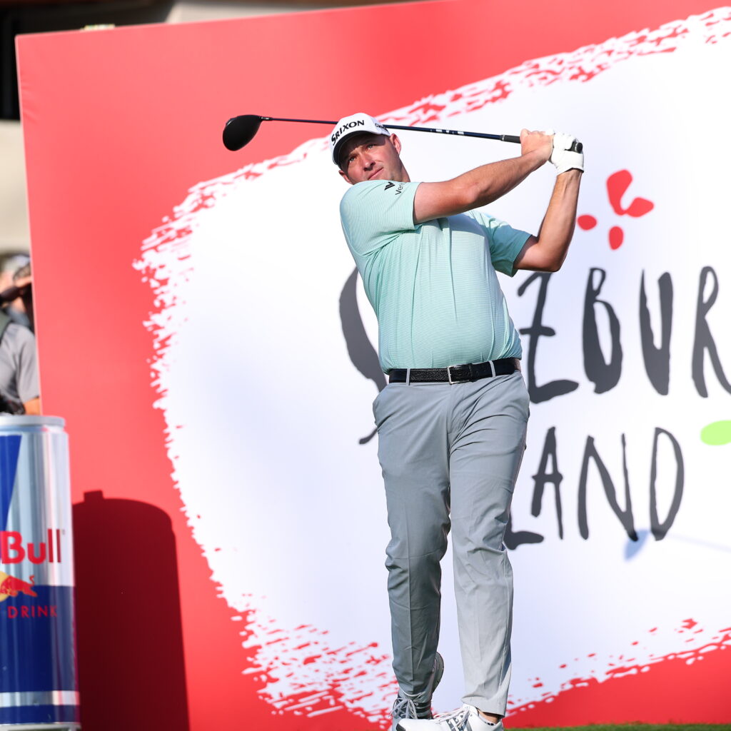 Ein Golfer schwingt einen Schläger auf einem Golfplatz, in dessen Nähe ein großes Cheeseburger Island-Schild und eine Red Bull-Dose stehen.