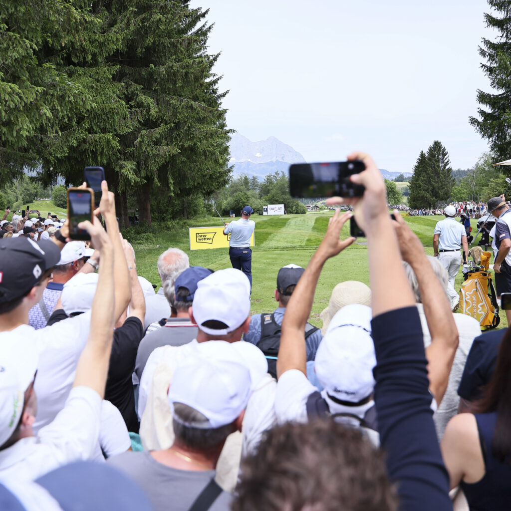 Eine Menschenmenge beobachtet und filmt einen Golfer, der bei einem Turnier abschlägt, umgeben von Bäumen und Bergen.