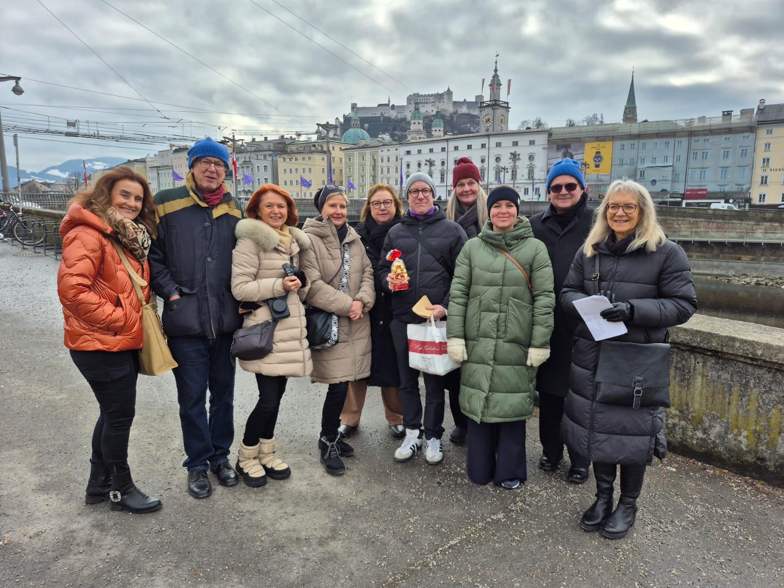 Eine Gruppe von Menschen in Winterkleidung posiert an einem Fluss mit einer Stadt und einem Schloss im Hintergrund.