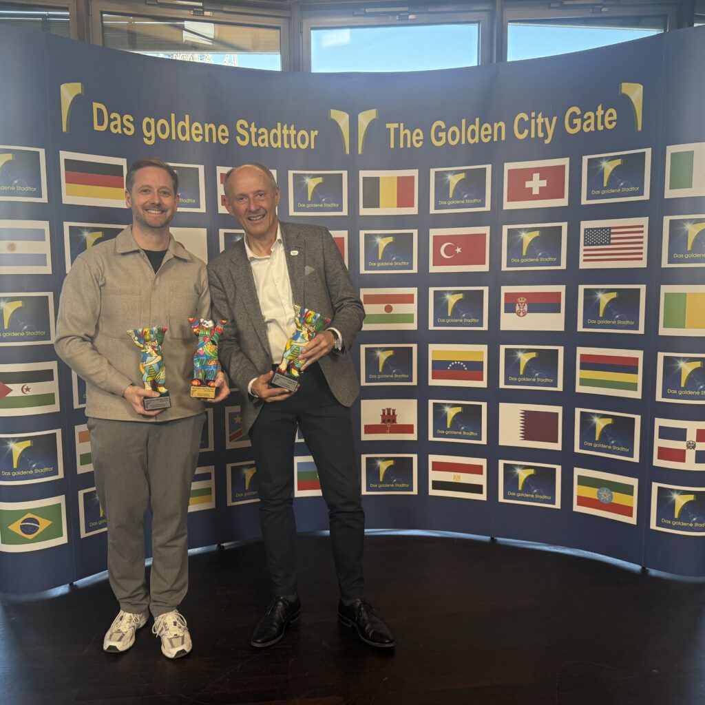 Zwei Männer, die bunte Trophäen in den Händen halten, stehen vor einer Kulisse mit internationalen Flaggen und der Aufschrift „The Golden City Gate“.