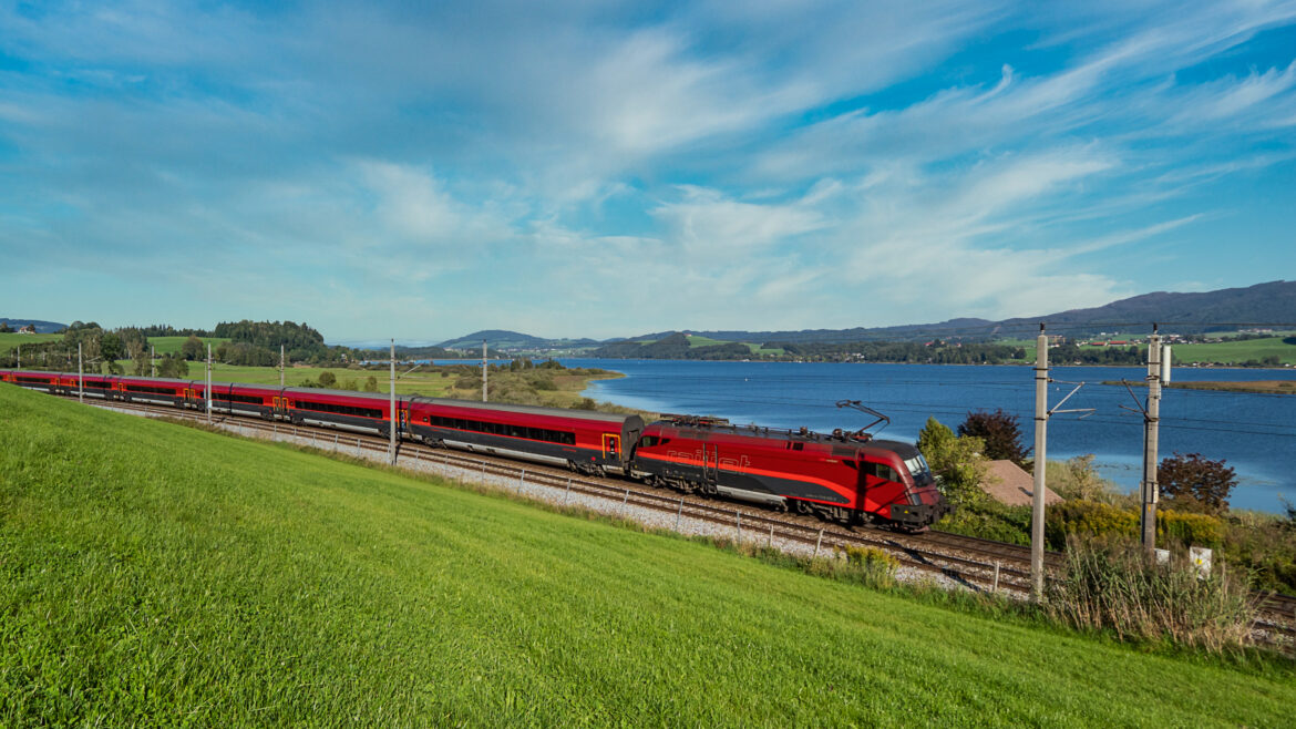 Ein roter Zug fährt auf Gleisen am Ufer eines Sees entlang, im Hintergrund sind grünes Gras und blauer Himmel zu sehen.