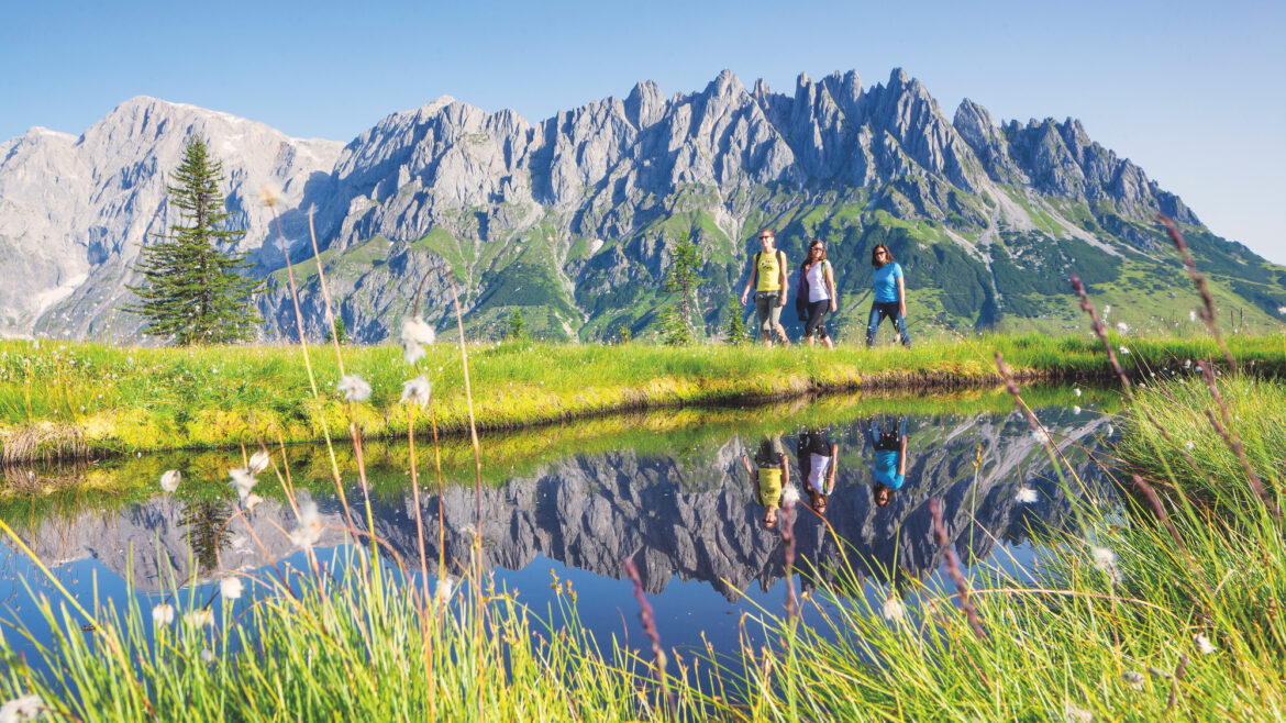 Vier Menschen spazieren auf einem grasbewachsenen Weg am Ufer eines Teiches entlang, während sich unter dem klaren Himmel die Berge im Wasser spiegeln.