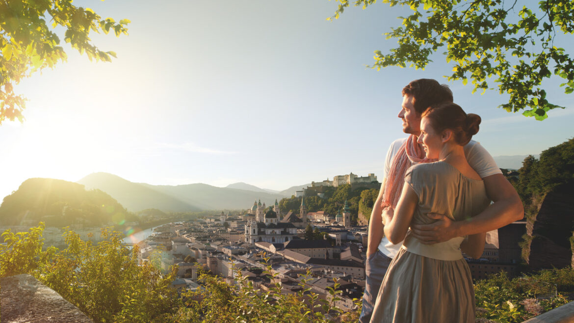 Ein Paar umarmt sich, während es bei Sonnenuntergang den Blick auf eine malerische Stadt genießt, umgeben von grünen Bäumen und Bergen.