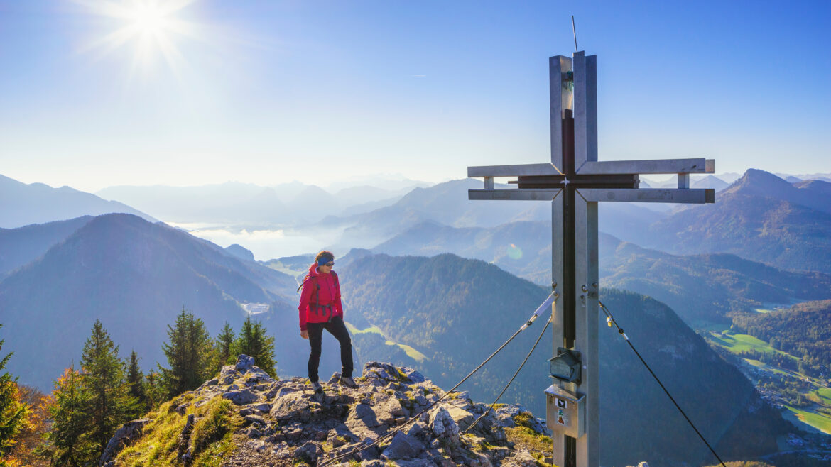 Ein Wanderer in roter Jacke steht neben einem Gipfelkreuz auf einem Berggipfel, im Hintergrund sind ein malerisches Tal und die Sonne zu sehen.