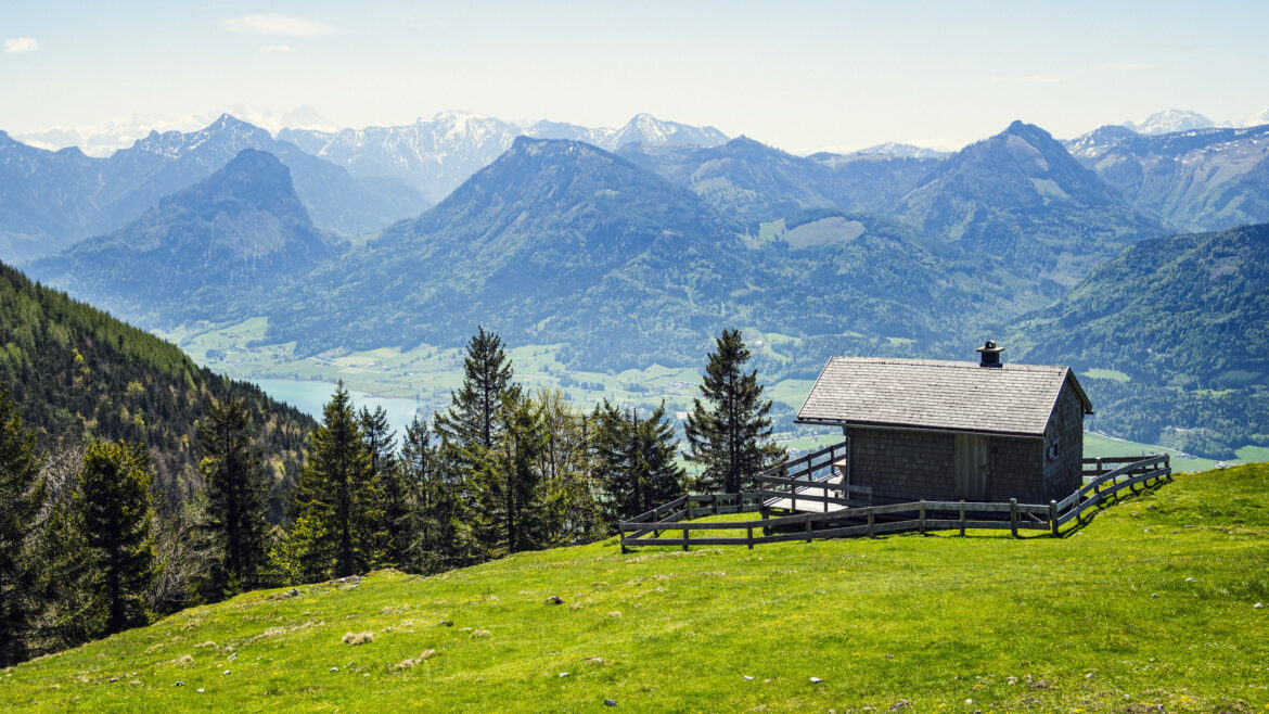 Eine kleine Hütte auf einem grasbewachsenen Hügel, im Hintergrund Berge, Bäume und ein See, über allem ein klarer Himmel.