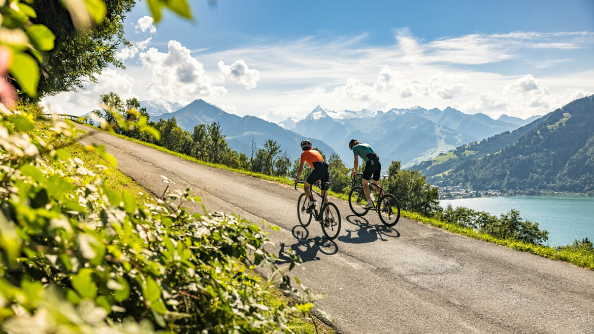 Zwei Radfahrer fahren bergauf auf einer malerischen Straße, im Hintergrund sind Berge, ein See und der blaue Himmel zu sehen.