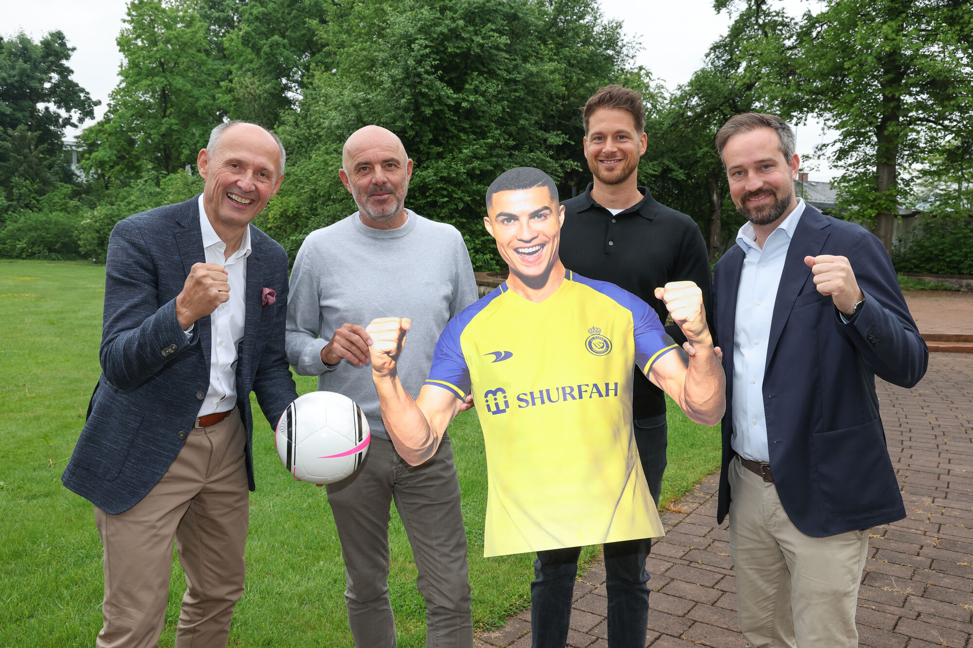 Vier Männer posieren im Freien mit einer Pappfigur eines Fußballspielers in einem gelben Trikot, der einen Fußball in der Hand hält.