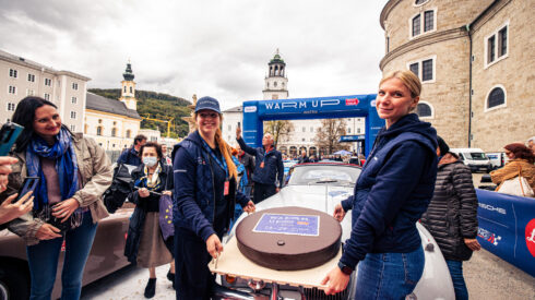 Zwei Frauen halten bei einer Veranstaltung im Freien vor einem Oldtimer ein großes rundes Schild hoch, während in der Nähe Zuschauer stehen.
