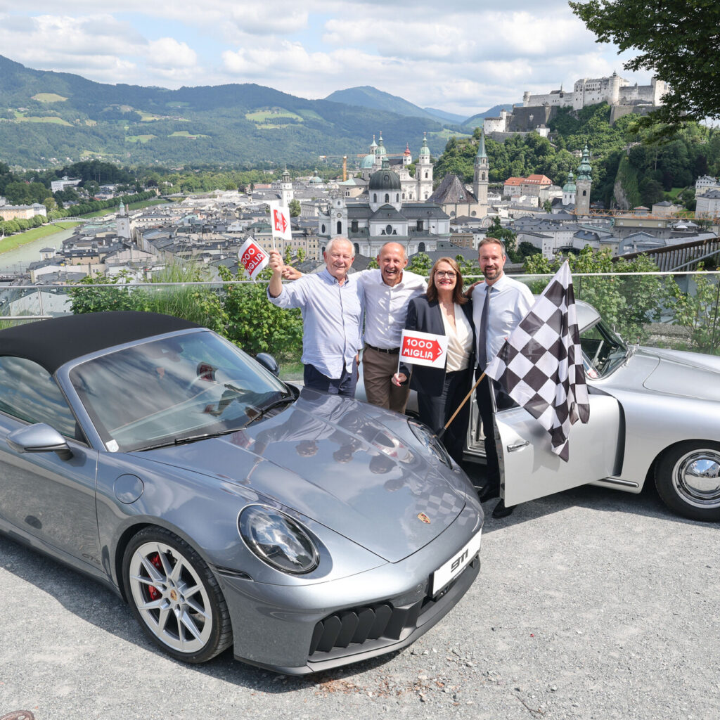 Fünf Menschen stehen lächelnd zwischen zwei silbernen Porsche-Fahrzeugen, im Hintergrund sind eine Stadt und Berge zu sehen.