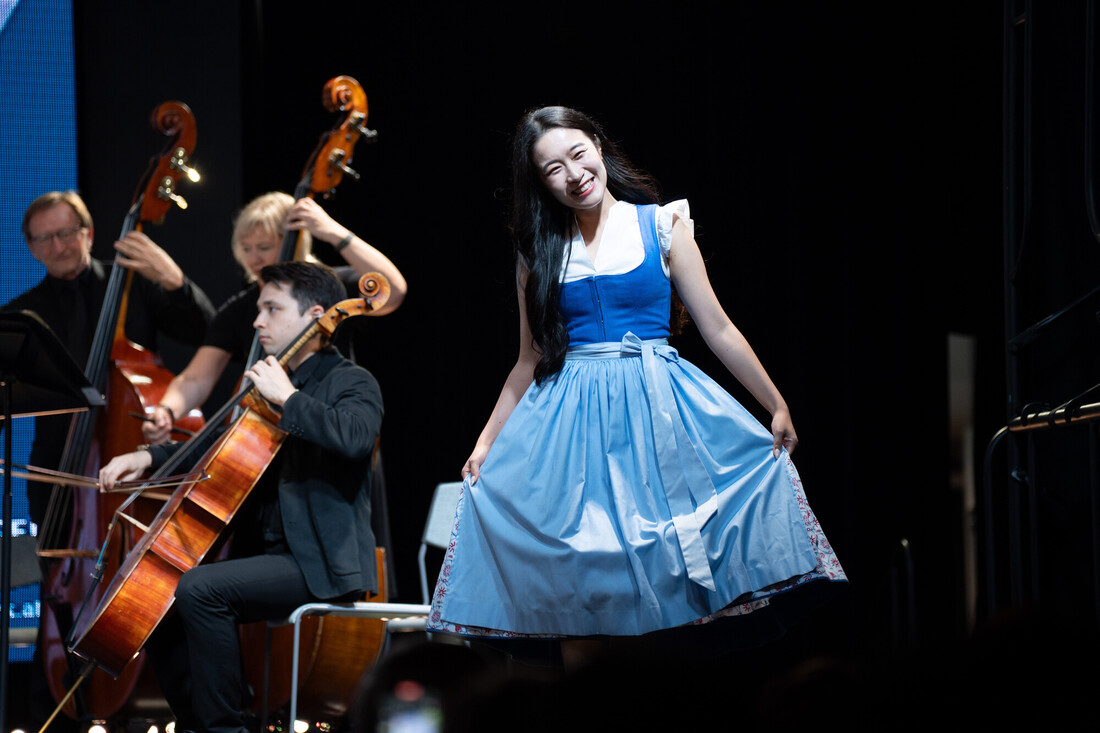 Eine Frau in einem blauen Kleid lächelt auf der Bühne vor Musikern, die Streichinstrumente spielen.
