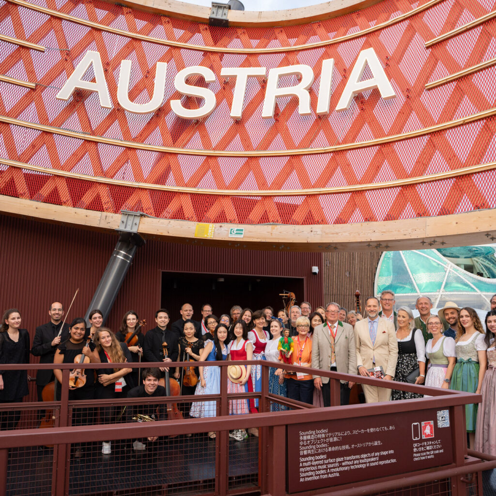Eine große Gruppe posiert bei einer Veranstaltung im Freien vor einem roten „AUSTRIA“-Schild; einige tragen traditionelle österreichische Trachten.