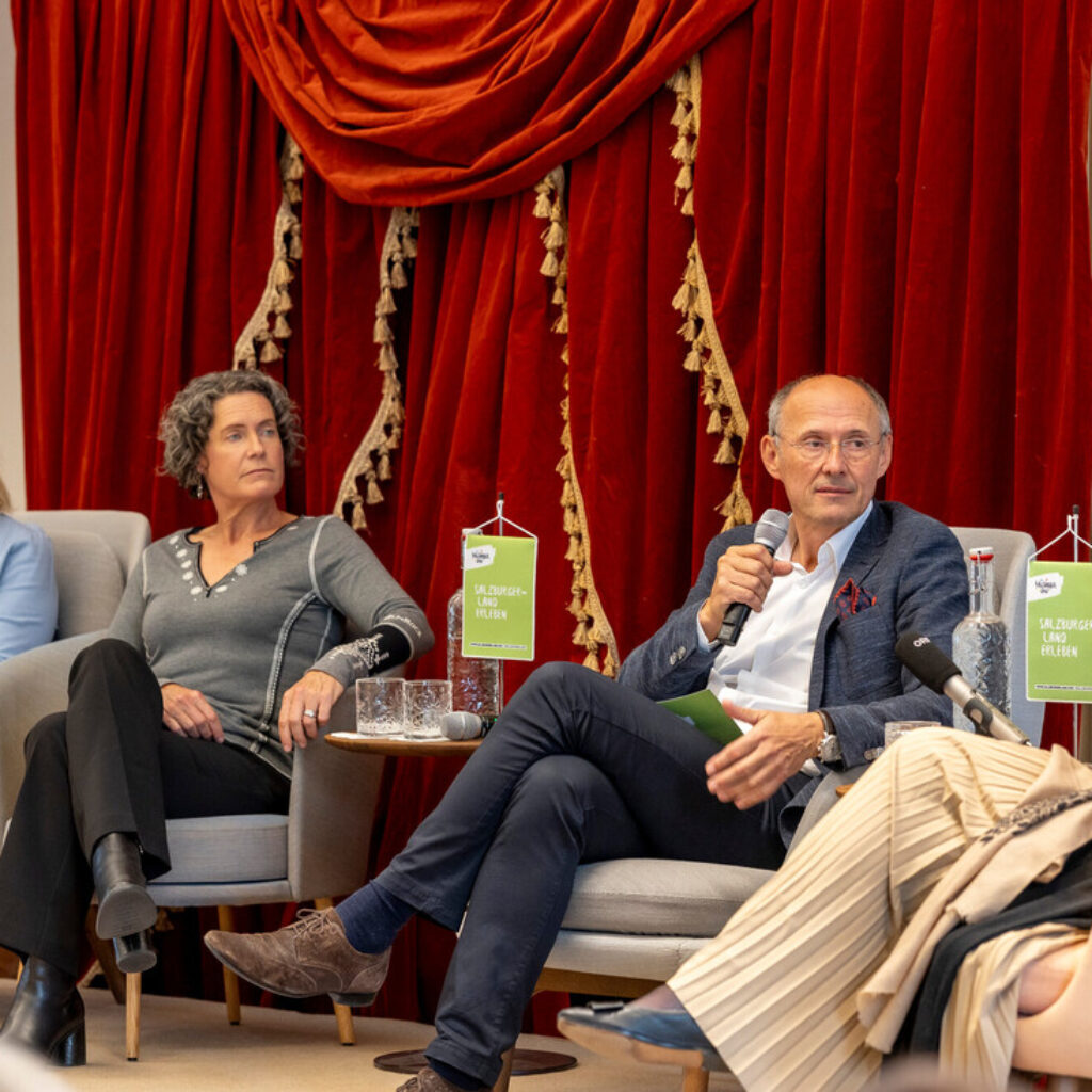 Vier Personen sitzen in Sesseln und führen eine Podiumsdiskussion vor einem roten Vorhang.