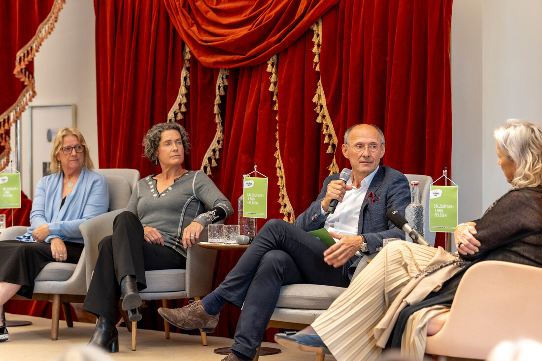 Vier Personen sitzen in Sesseln und führen eine Podiumsdiskussion vor einem roten Vorhang.