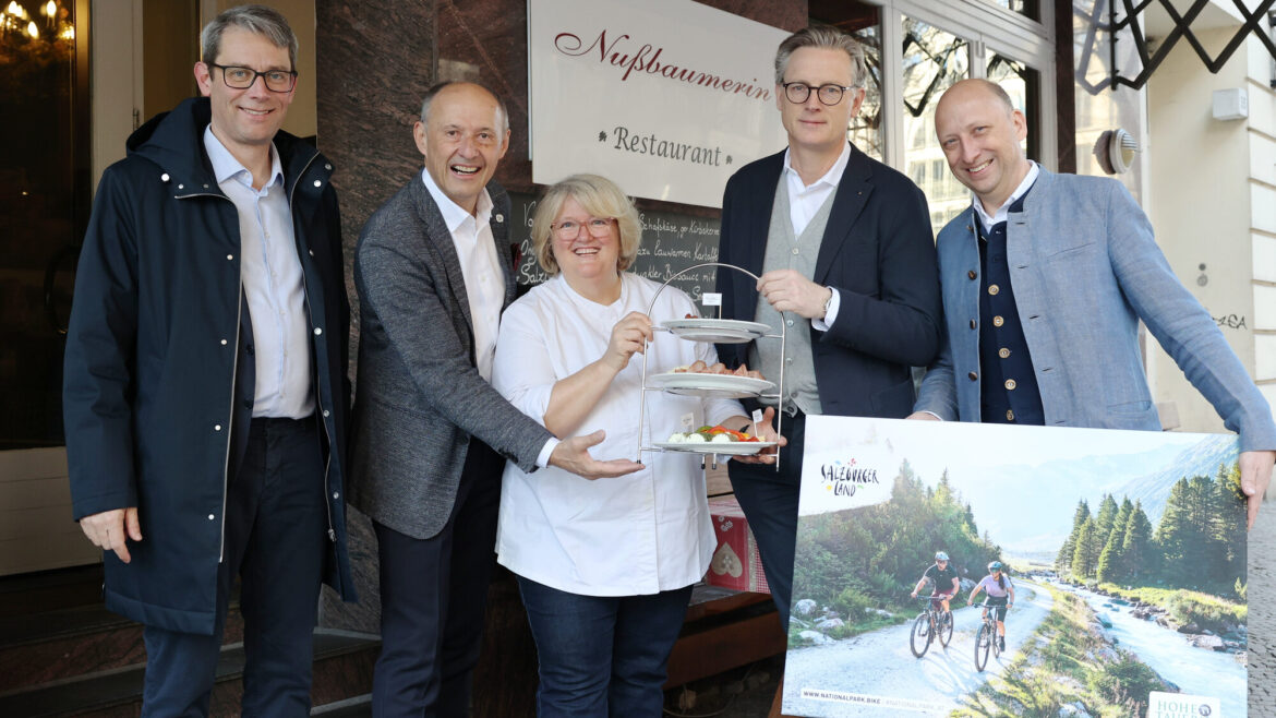 Fünf Menschen stehen lächelnd vor einem Restaurant; einer hält ein Tablett mit Essen, ein anderer ein Plakat mit einem Fahrradmotiv in der Hand.