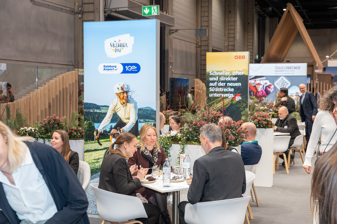 Menschen, die an Tischen in einer belebten Kongresshalle Kontakte knüpfen und sich unterhalten, mit Werbedisplays im Hintergrund.