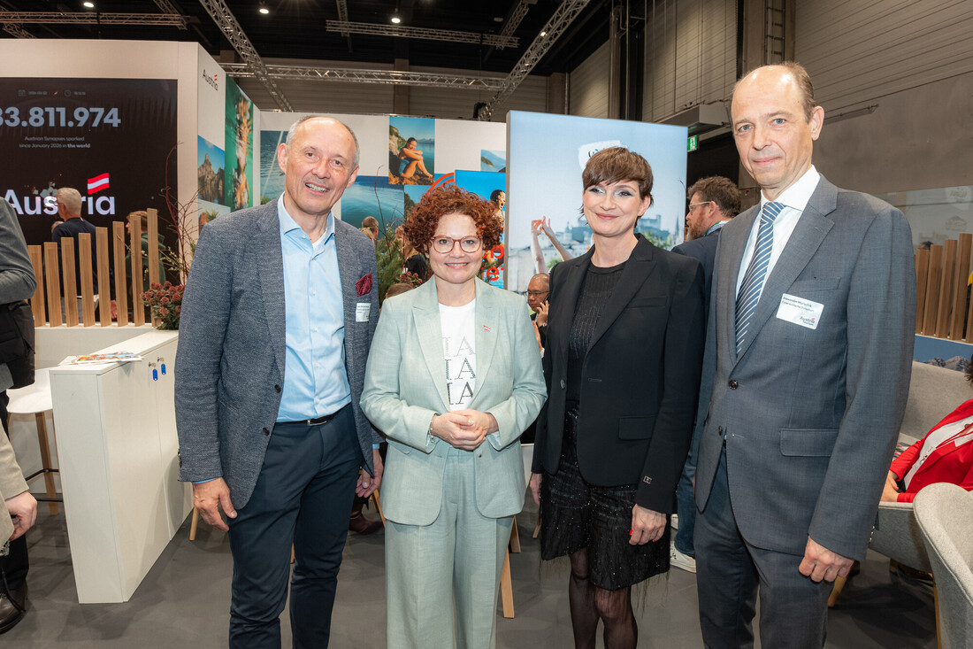 Vier professionell gekleidete Personen stehen lächelnd bei einer Veranstaltung oder Konferenz in einer Halle mit Ständen hinter ihnen zusammen.