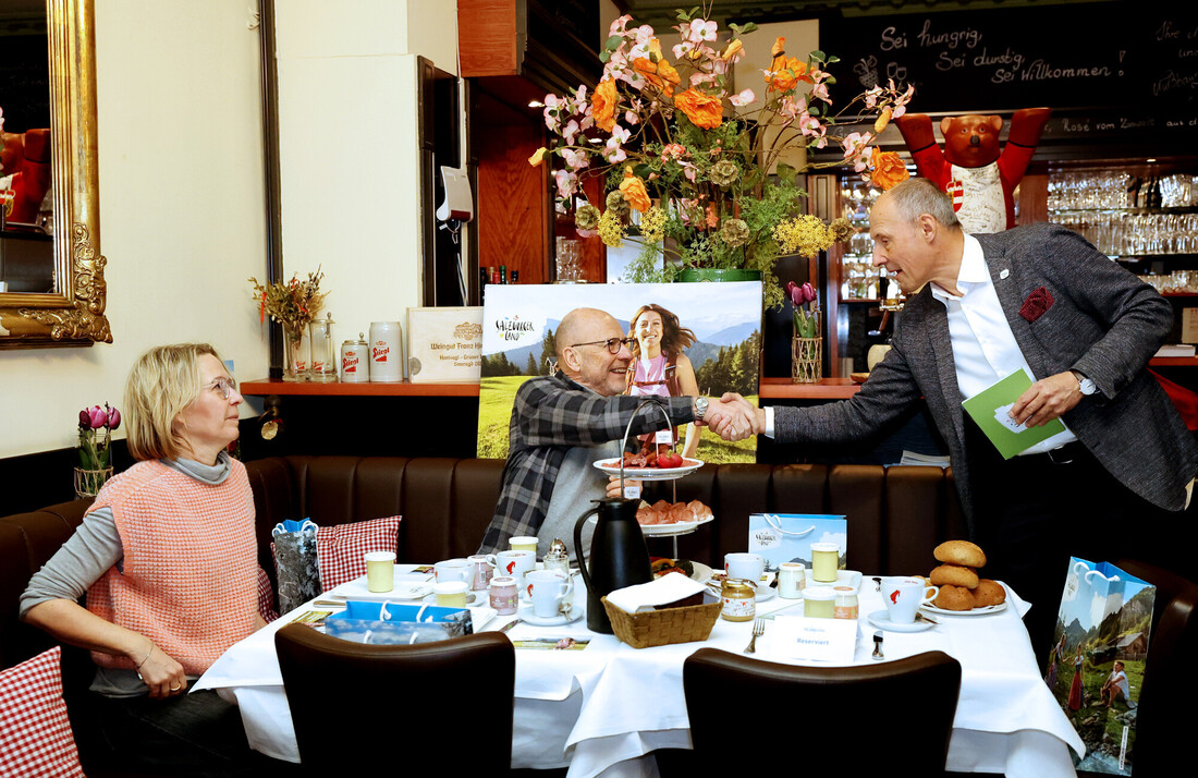 Drei Menschen treffen sich in einem gemütlichen Café, schütteln sich die Hände an einem für das Frühstück gedeckten Tisch, Blumen im Hintergrund.