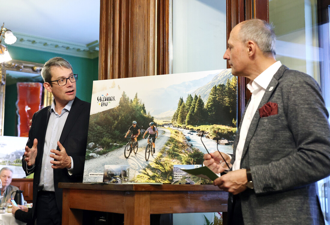 Zwei Männer in Anzügen diskutieren neben einem großen Foto von Radfahrern, die auf einem malerischen Bergweg in der Nähe eines Flusses fahren.