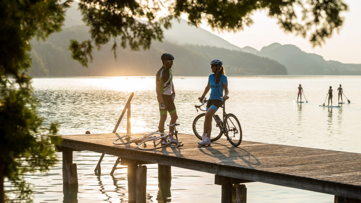 Zwei Radfahrer unterhalten sich bei Sonnenuntergang auf einem Steg an einem See, im Hintergrund sind Paddleboarder zu sehen.